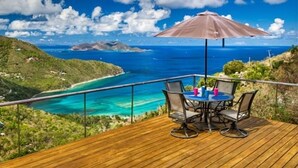 Una terraza de madera con mesa y sillas, con vista al mar turquesa y colinas verdes.