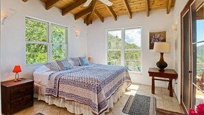 Un dormitorio con techo de madera, una cama con colcha estampada, dos ventanas, una mesita de noche con lámpara y vistas al exterior con vegetación.