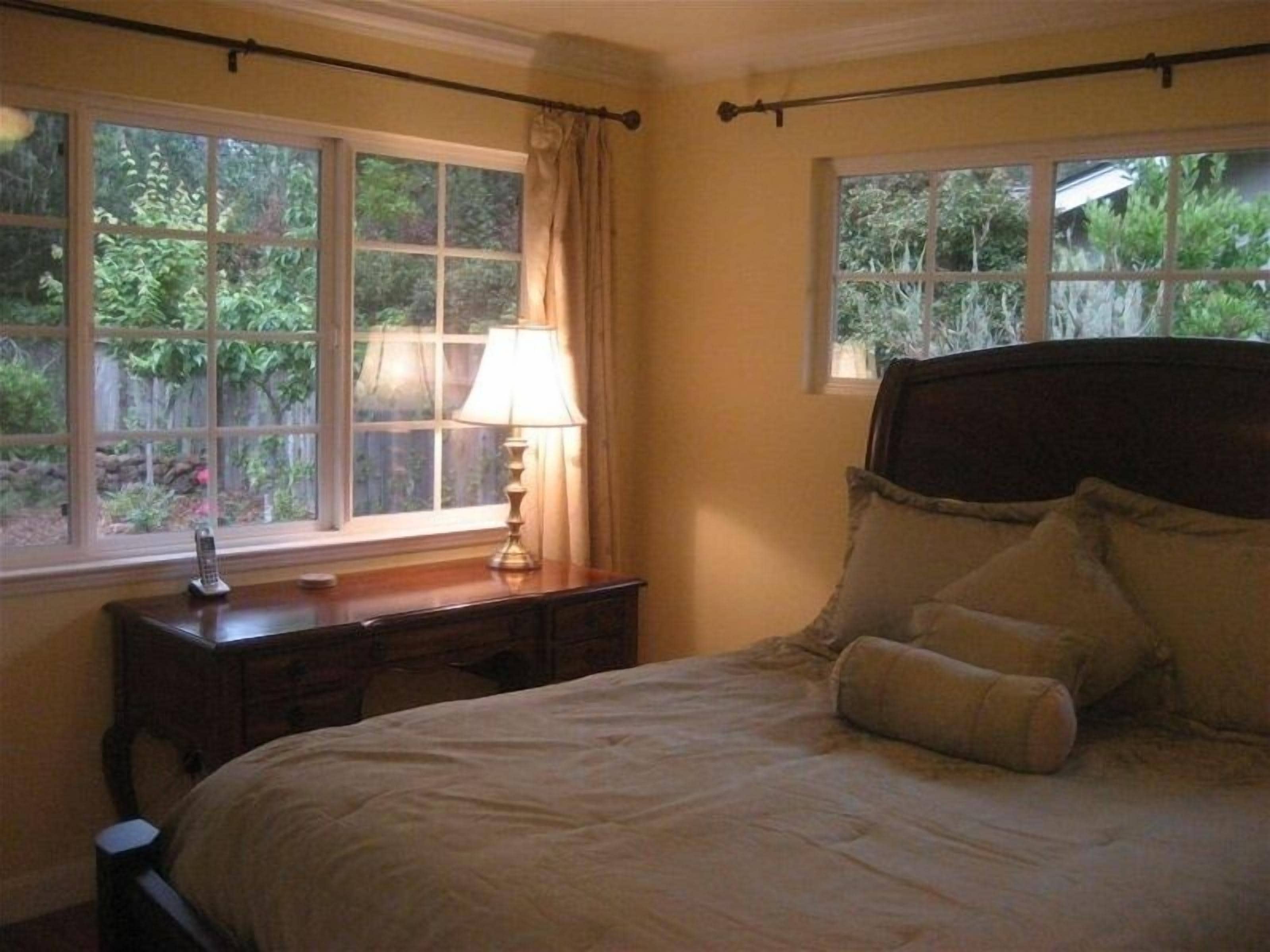 Une chambre à coucher avec un lit, une table de chevet, une lampe et deux fenêtres avec des rideaux.