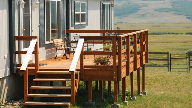 Una terraza de madera con una silla y una mesa, una escalera que lleva a una casa con contraventanas, y vistas a un campo cubierto de hierba.