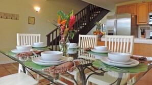 A dining area with a glass table set for four, surrounded by white chairs. A staircase is visible in the background leading to a kitchen area with wooden cabinets.