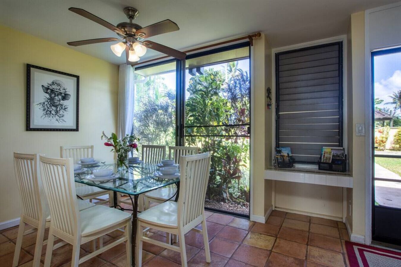 Un comedor con una mesa de vidrio, sillas blancas, un ventilador de techo y vistas al exterior a zonas verdes.