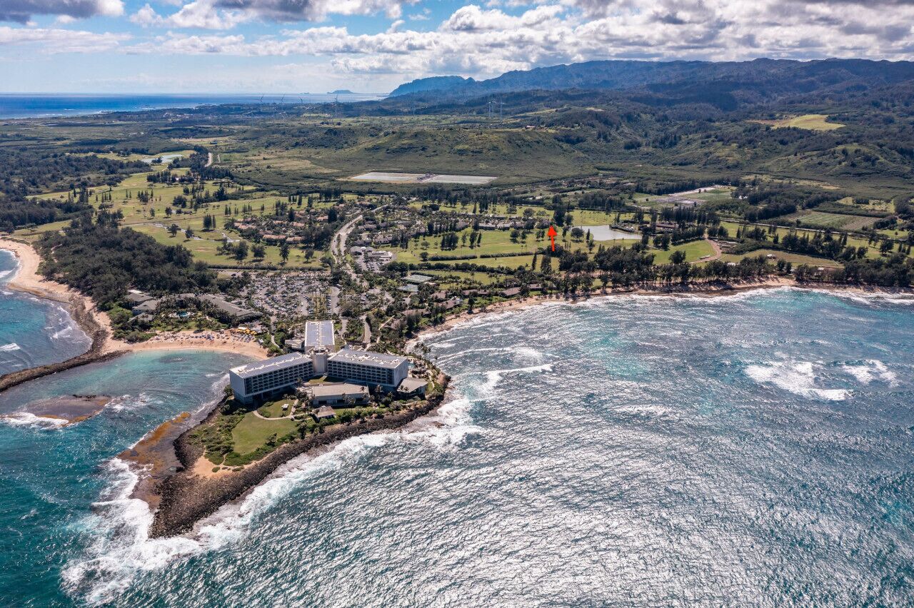 An aerial view of a coastal resort with a large building near the beach, surrounded by lush greenery and hilly terrain.