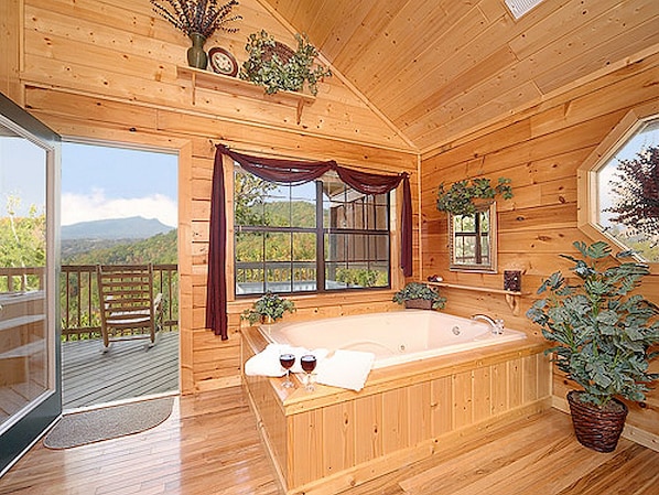 Une cabane en bois dotée d’une grande baignoire en bois, d’un balcon offrant une vue sur les montagnes et d’une grande fenêtre avec des rideaux.