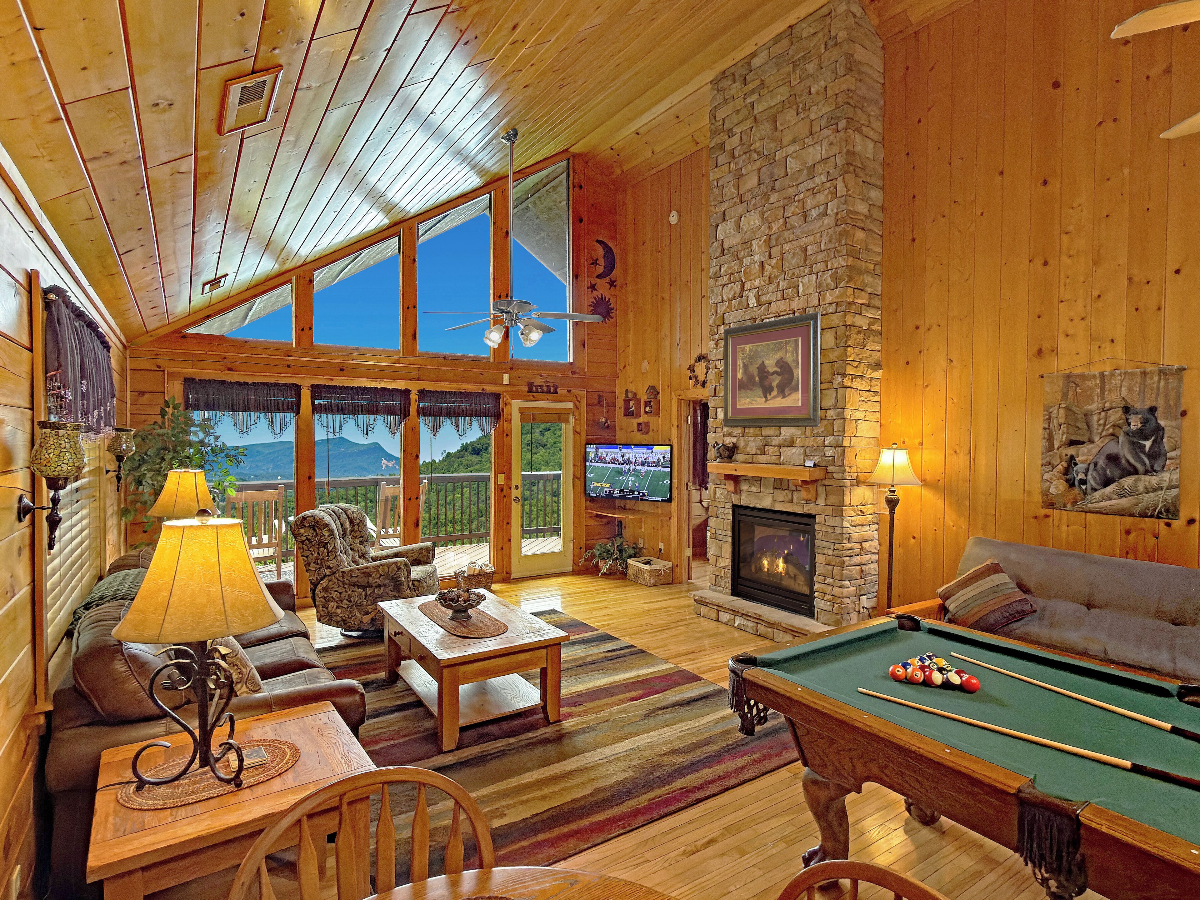 Interior acogedor de una cabaña con mesa de billar, asientos cómodos, chimenea y un amplio ventanal con vistas a la montaña.