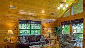 Ein gemütliches Blockhaus-Interieur mit Holzbalkendecke, einer Couch, einem Sessel, einem Couchtisch und einem großen Fenster mit Blick ins Grüne.