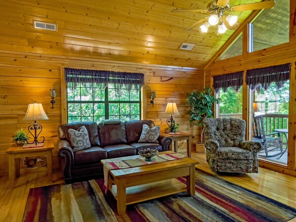 A cozy cabin interior with a wooden ceiling, a sofa, a chair, a coffee table, and a large window offering a view of greenery outside.