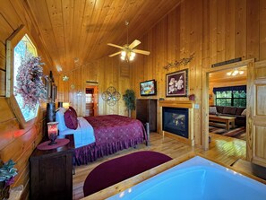 A wooden cabin interior with a bed, a fireplace, a television, and a view of the outdoors.