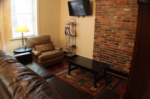 A living room with a leather couch, a small table, a bookshelf, and a brick wall. - Cozy Fells Point Gem / In The Heart of Fells Point (Baltimore)