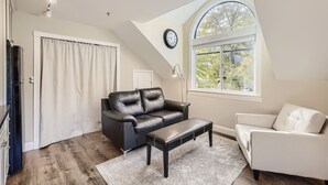 A living room with a black leather sofa, a white armchair, and a matching ottoman. A large window provides natural light, and a clock is visible on the wall. - Downtown Boulder Guest House for 2 guests (Boulder)