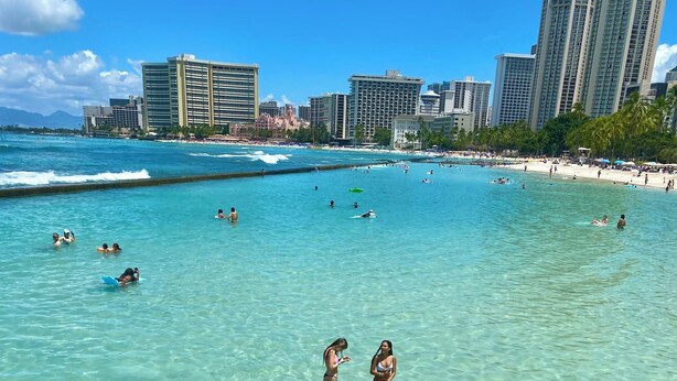 Une plage à l’eau turquoise et limpide, où l’on peut voir des gens nager et faire de la planche à voile, et un paysage urbain avec de hauts immeubles en arrière-plan.