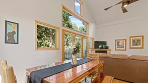 Ein Wohnzimmer mit Esstisch aus Holz, einer Couch und einem Fernseher. Große Fenster bieten Blick auf Bäume.