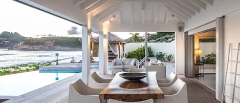 Ein moderner Essbereich im Freien mit einem Holztisch und weißen Stühlen, mit Blick auf einen Pool und eine schöne Küstenlandschaft.