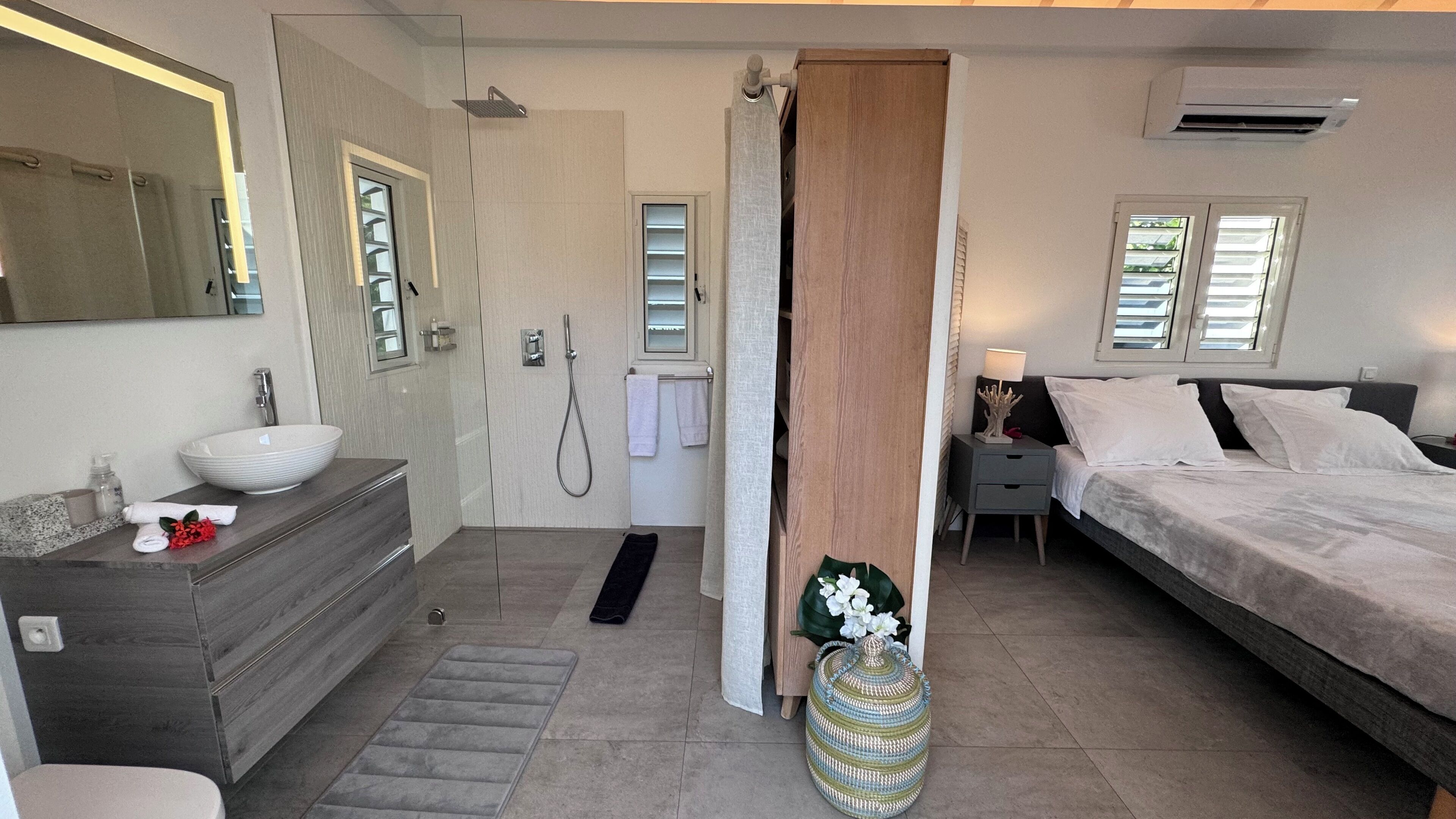 A modern bedroom with a large bed, a bedside table with a lamp, a bathroom with a sink and shower, and a large mirror.