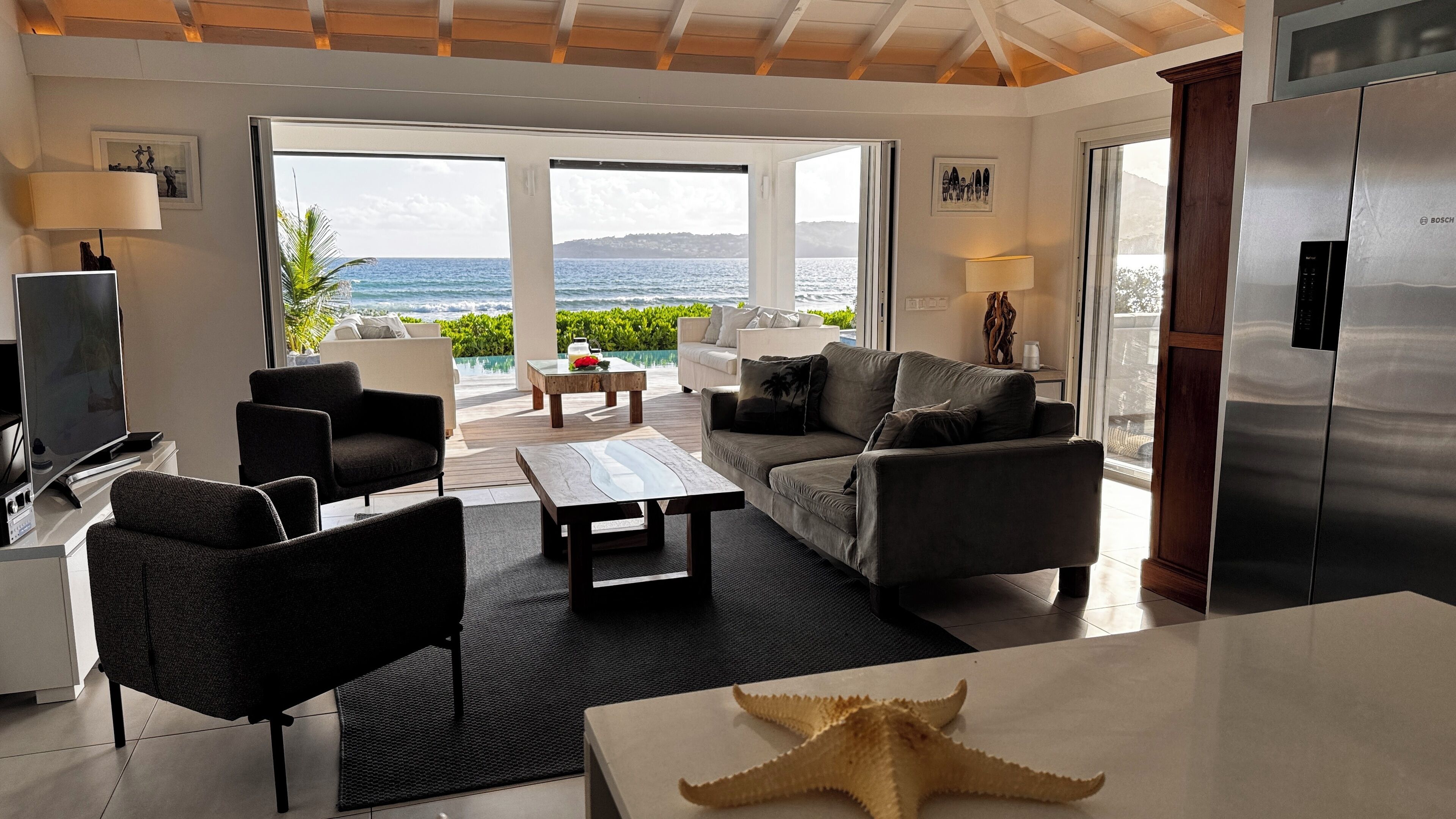 A modern living room with a large window overlooking a pool and ocean, furnished with a sofa, chairs, and a coffee table.