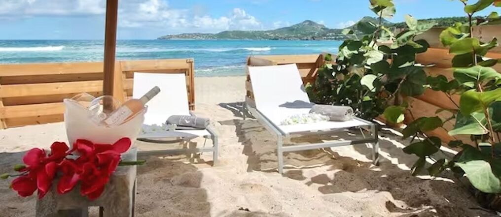 A beachside lounge with two white sun loungers, a bucket with a bottle and flowers, and a wooden fence with greenery.