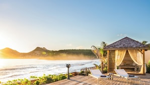 Un resort frente a la playa con un pabellón de madera, sillas de descanso y vistas al océano y a las montañas.