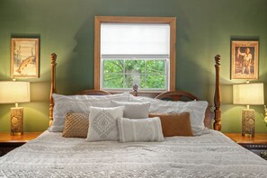 A neatly made bed with a patterned bedspread and multiple pillows, a window with a view of greenery, and two bedside tables with lamps.