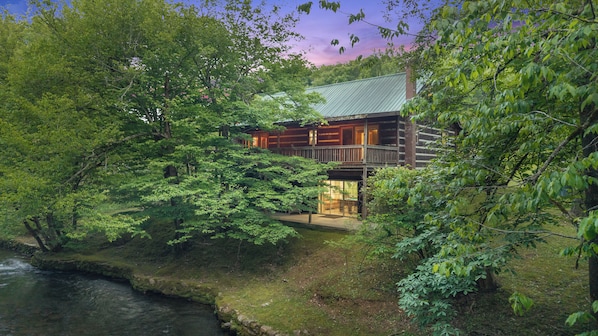 A log cabin with a deck, surrounded by dense forest and a stream.