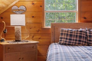 A wooden cabin room with a bed, a nightstand with a lamp, a heart-shaped mirror, and a window with a view of trees. - STUNNING CABIN, TROUT STREAM, HUGE GAMEROOM, HOT TUB,FIRE PIT,TUBING,& MEMORIES! (Cosby)