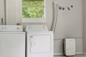 A laundry area with two washing machines, a dryer, and a window with a view of trees. - STUNNING CABIN, TROUT STREAM, HUGE GAMEROOM, HOT TUB,FIRE PIT,TUBING,& MEMORIES! (Cosby)