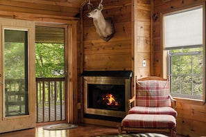 A wooden cabin interior with a fireplace, a plaid chair, and a deer head mounted on the wall.