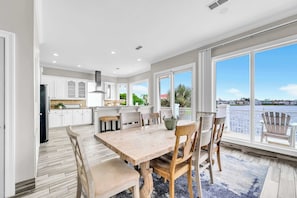 A spacious kitchen with white cabinets, a wooden dining table, and large windows overlooking a body of water.