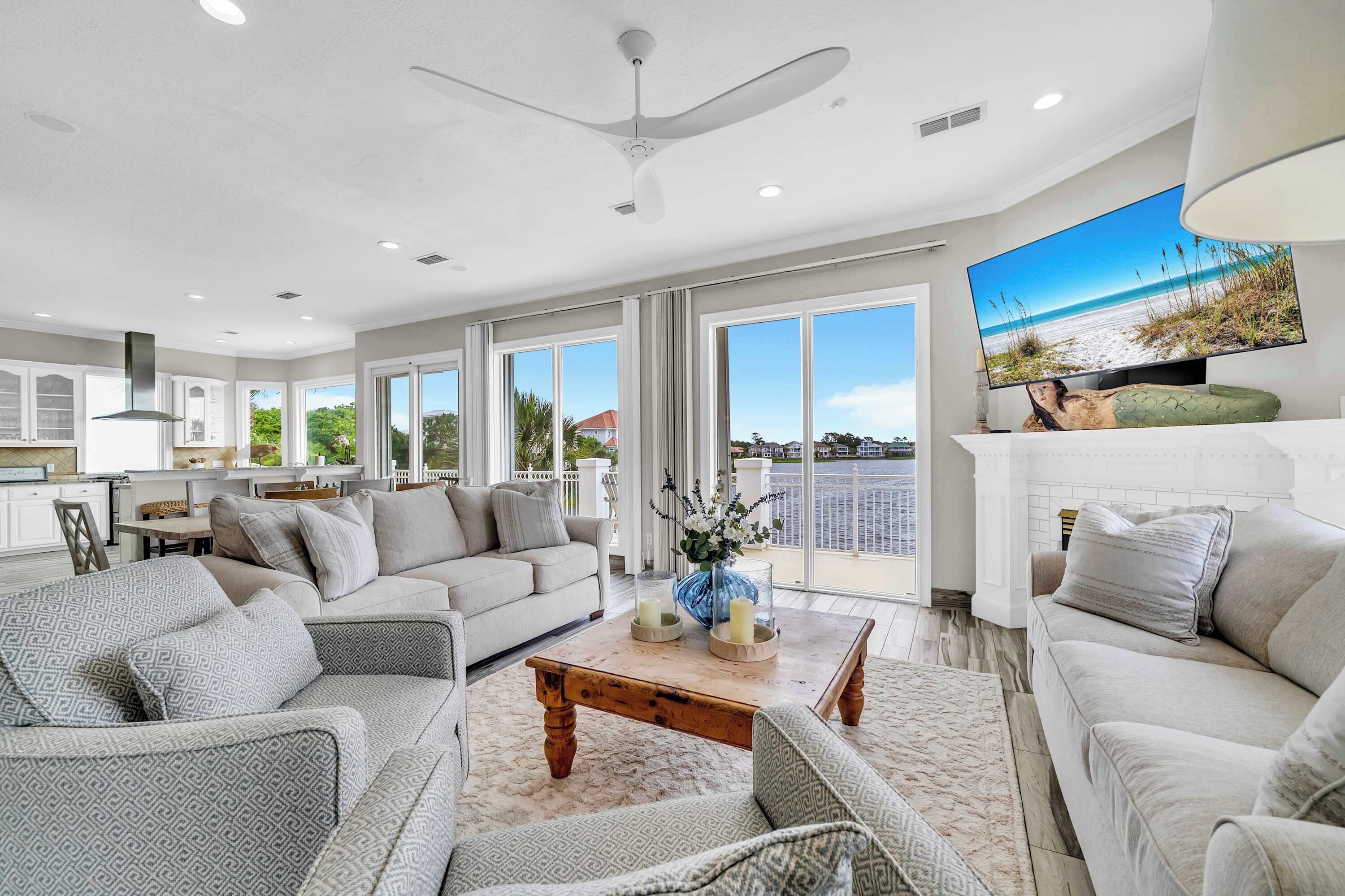 A spacious living area with a large flat-screen TV, a ceiling fan, and a balcony with a view of the ocean.