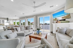 A spacious living area with a large flat-screen TV, a ceiling fan, and a balcony with a view of the ocean.