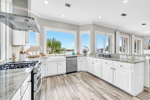 A modern kitchen with white cabinets, a stainless steel range hood, and a large window offering a view of palm trees.
