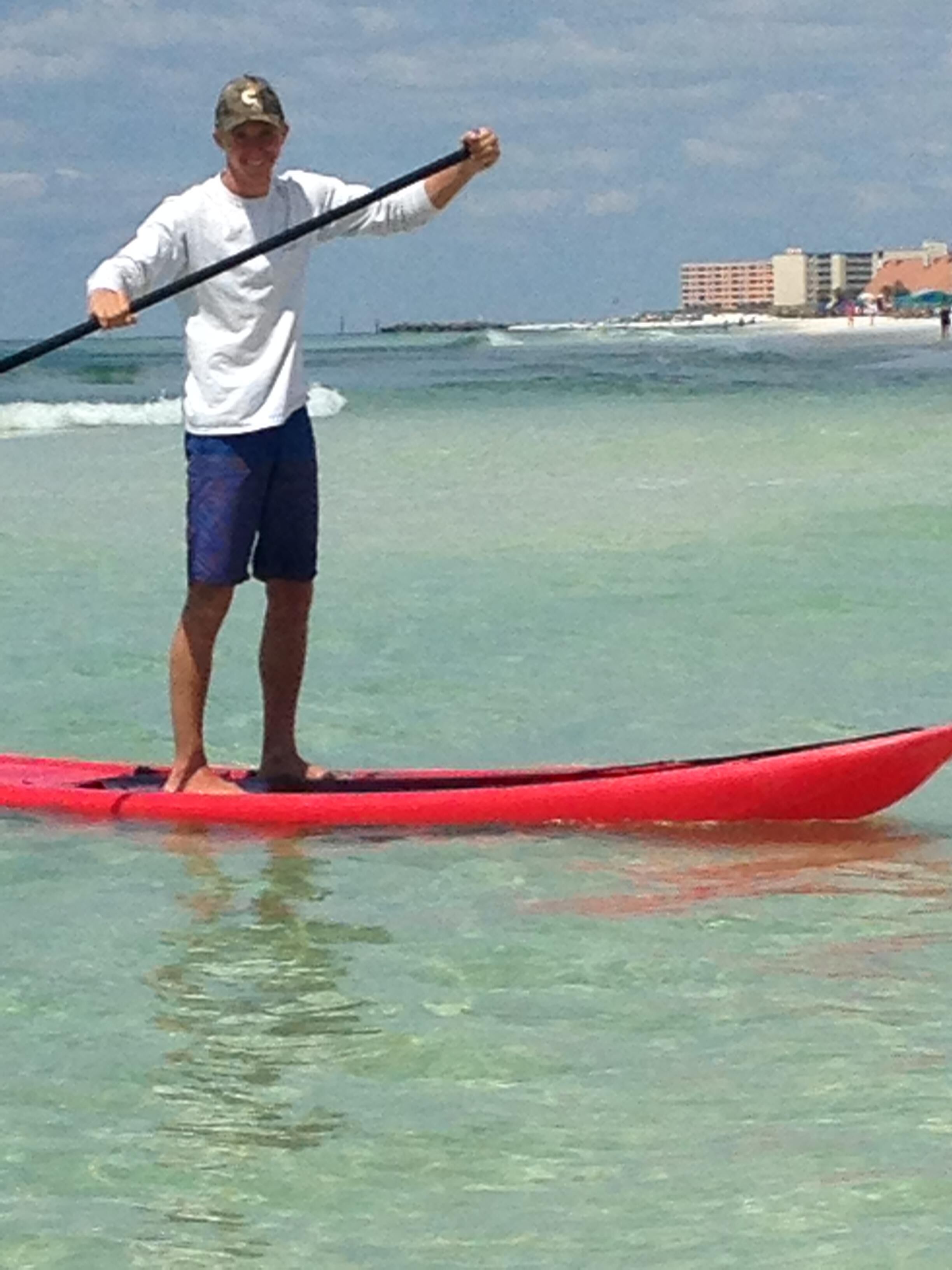 Una persona in piedi su una tavola da paddleboard rossa in acque limpide, con edifici sullo sfondo.