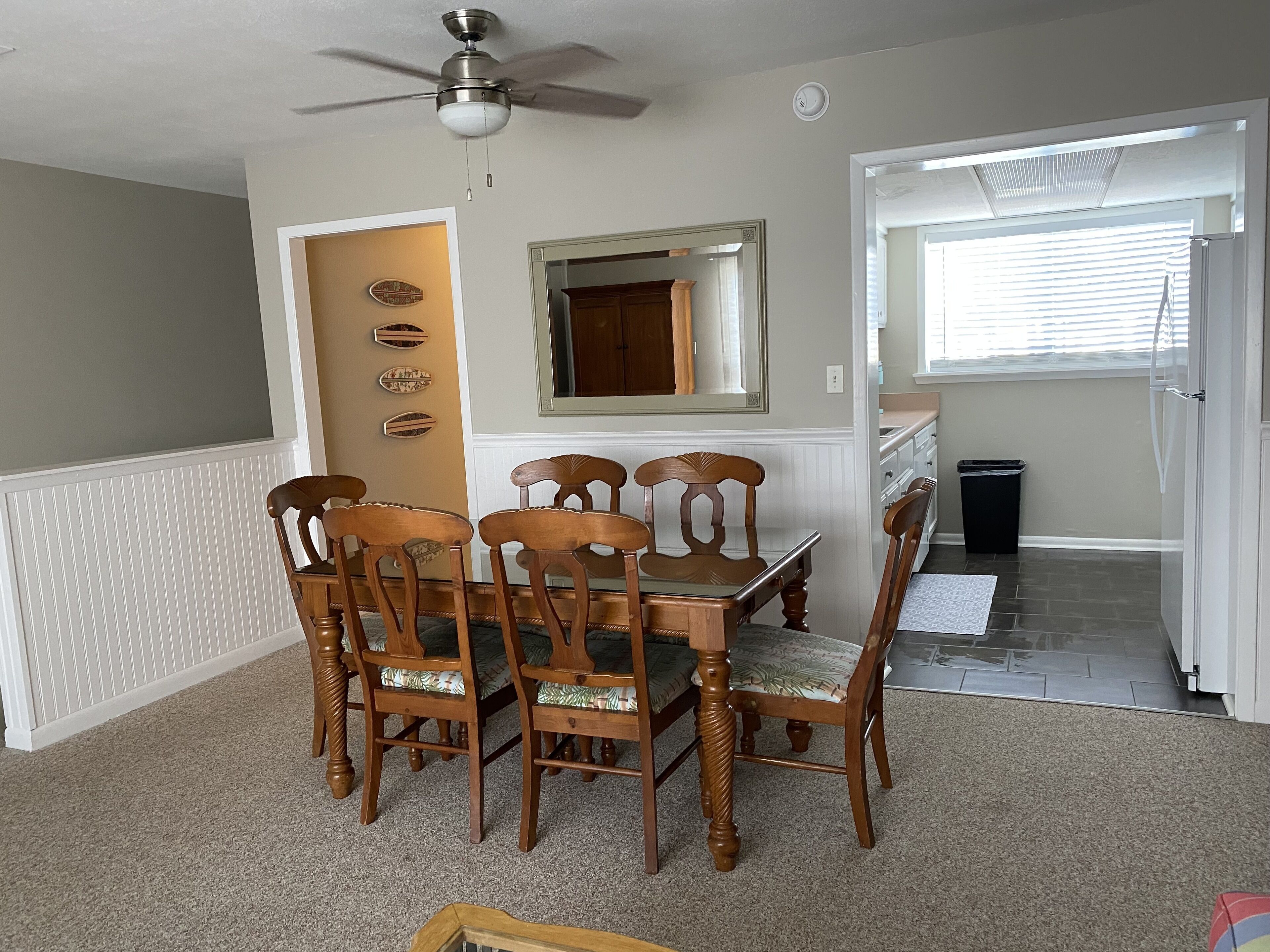 A dining area with a wooden table and chairs, a ceiling fan, a mirror, and a door leading to a kitchen.