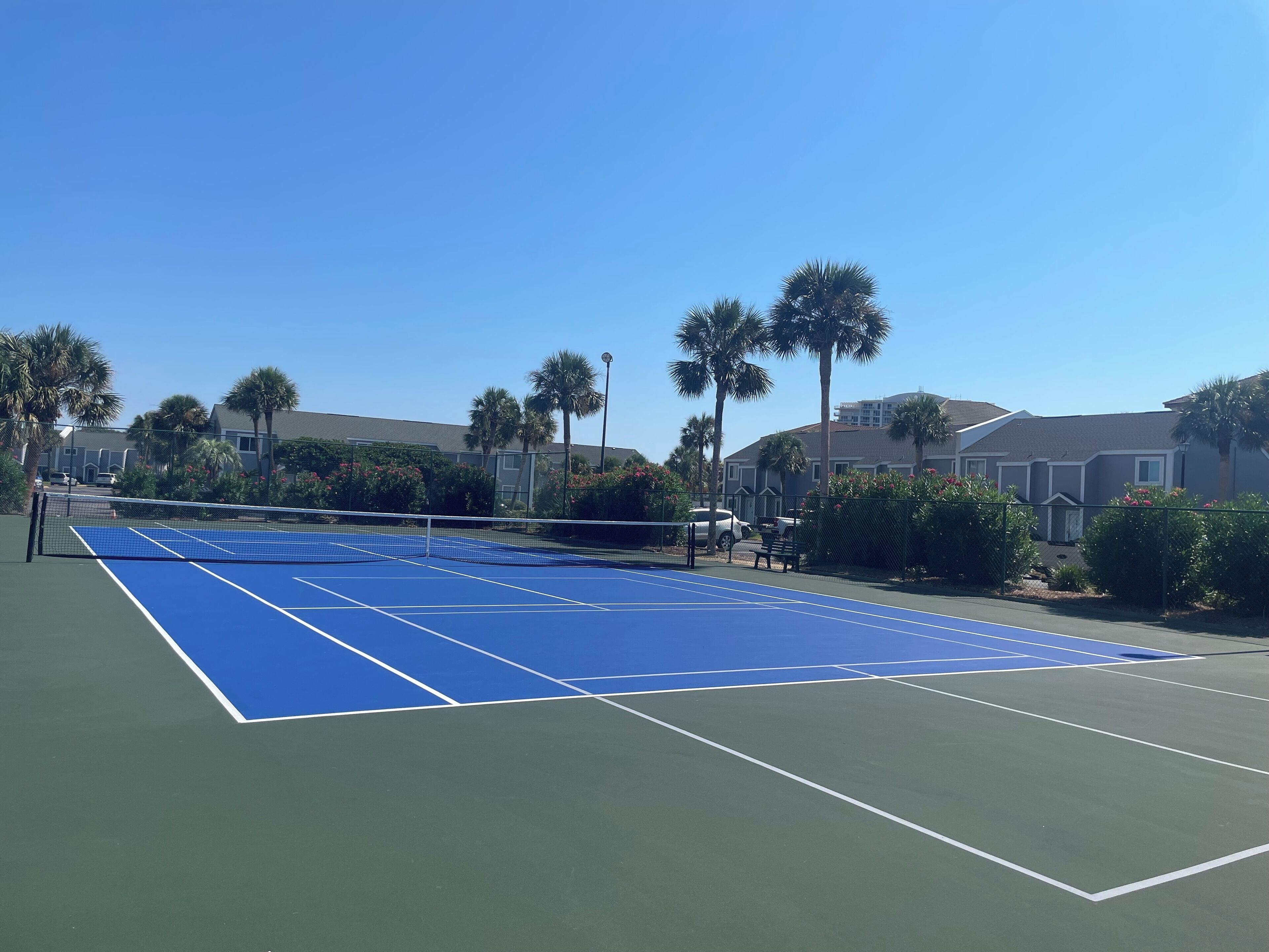 Una cancha de tenis al aire libre con superficie azul, rodeada de palmeras y edificios residenciales.