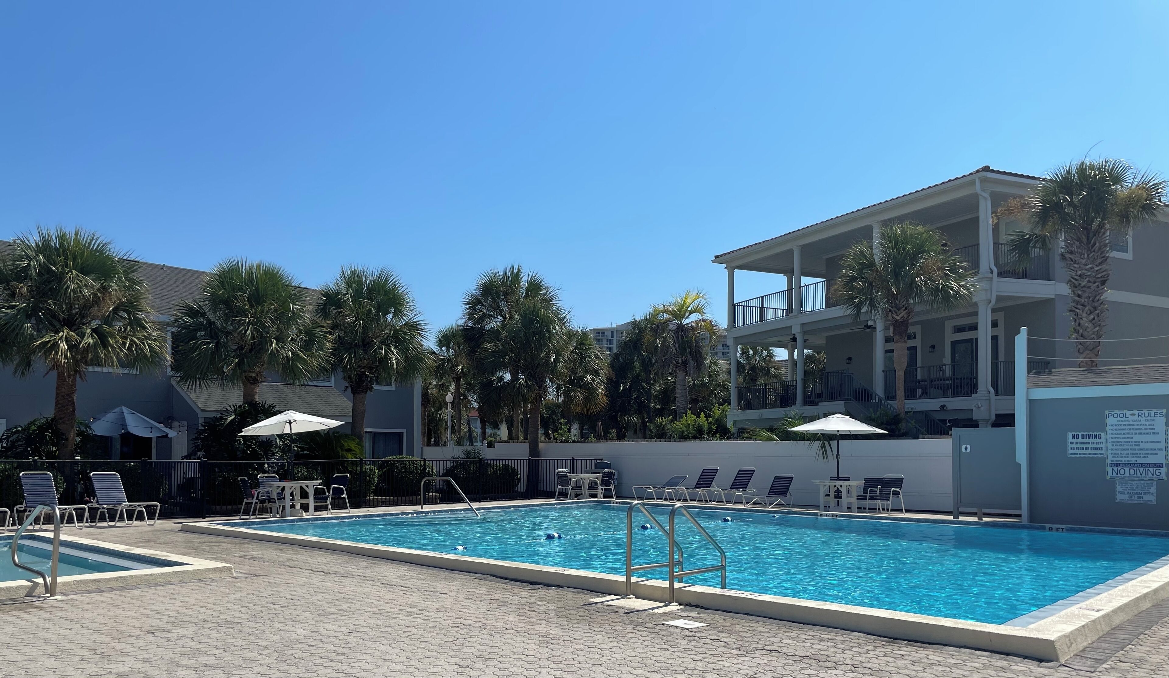 A swimming pool with lounge chairs and umbrellas, surrounded by palm trees and a multi-story building.
