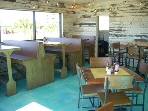 Un intérieur rustique avec des tables et des bancs en bois, un sol bleu et une vue sur les palmiers à l’extérieur.