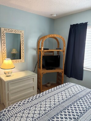 Une chambre à coucher comprenant un lit, une commode, un miroir, un téléviseur posé sur un meuble et une fenêtre avec des rideaux.