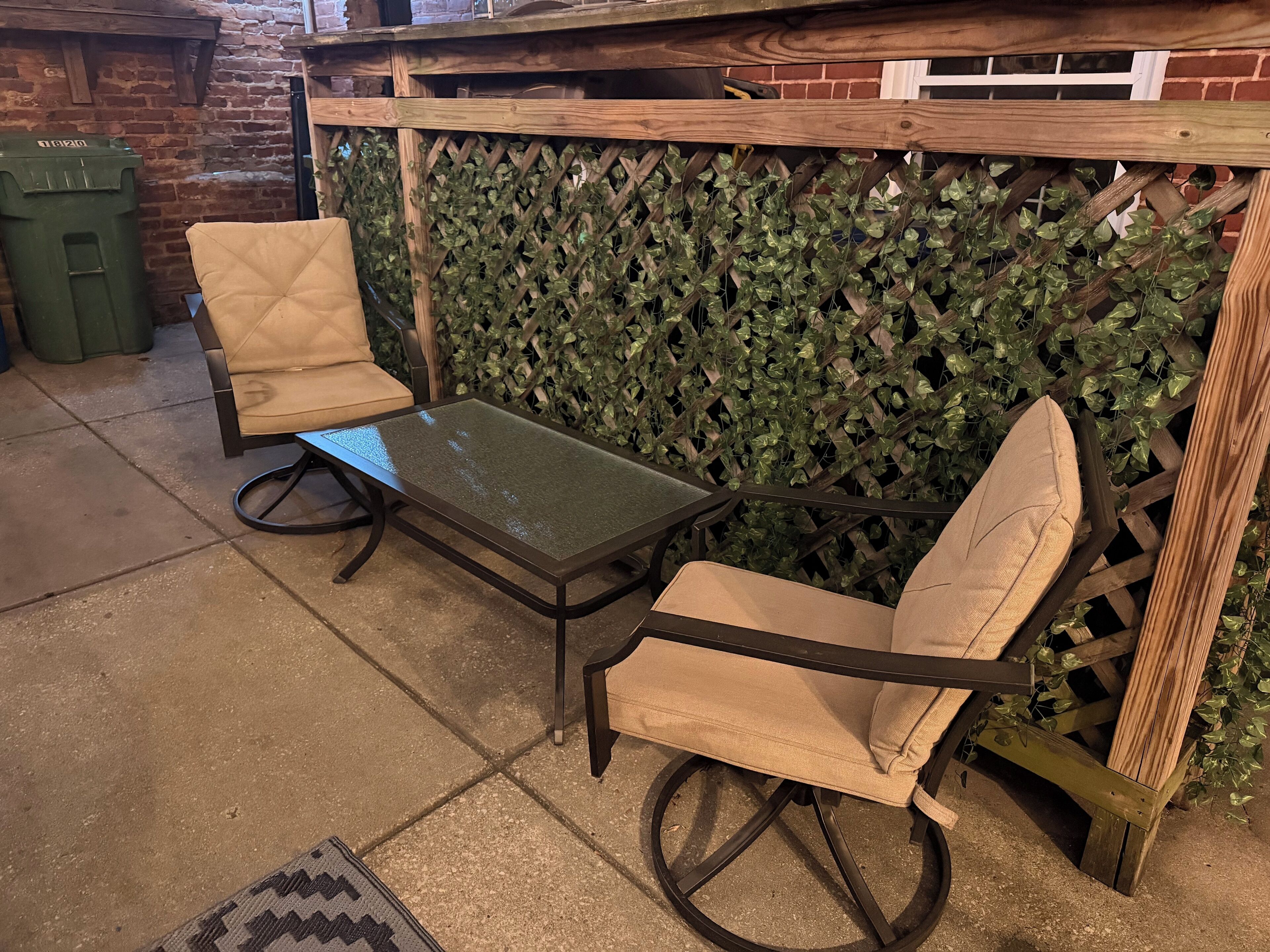 A patio area with a table and two chairs, a brick wall, and a green trash bin.
