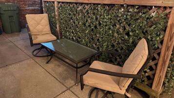 A patio area with a table and two chairs, a brick wall, and a green trash bin.