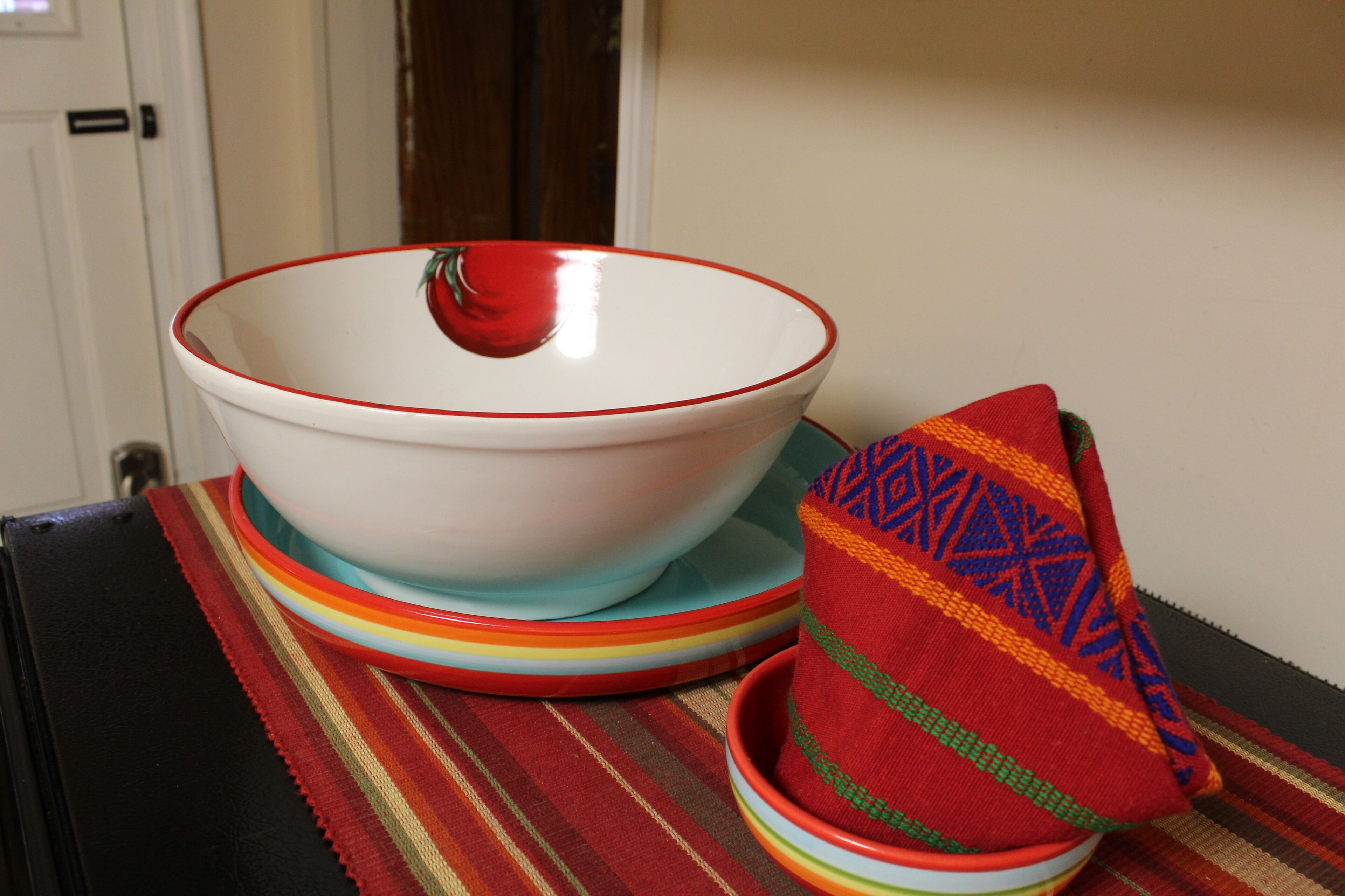 A white bowl with a red rim, a colorful striped cloth, and a red and green patterned napkin on a table.