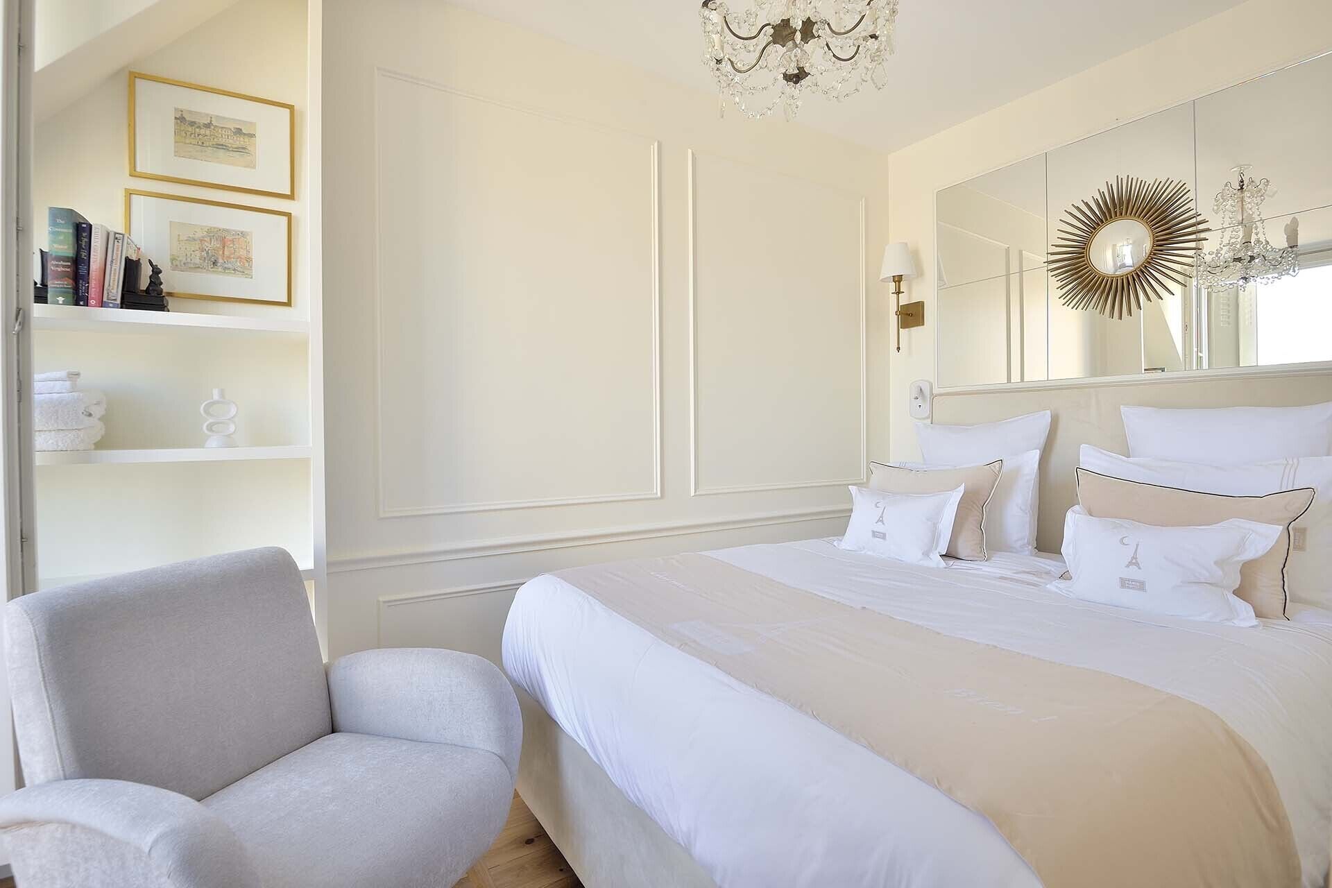 A bedroom with a large bed, a chair, a bookshelf with books and framed pictures, and a mirror with a sunburst design.