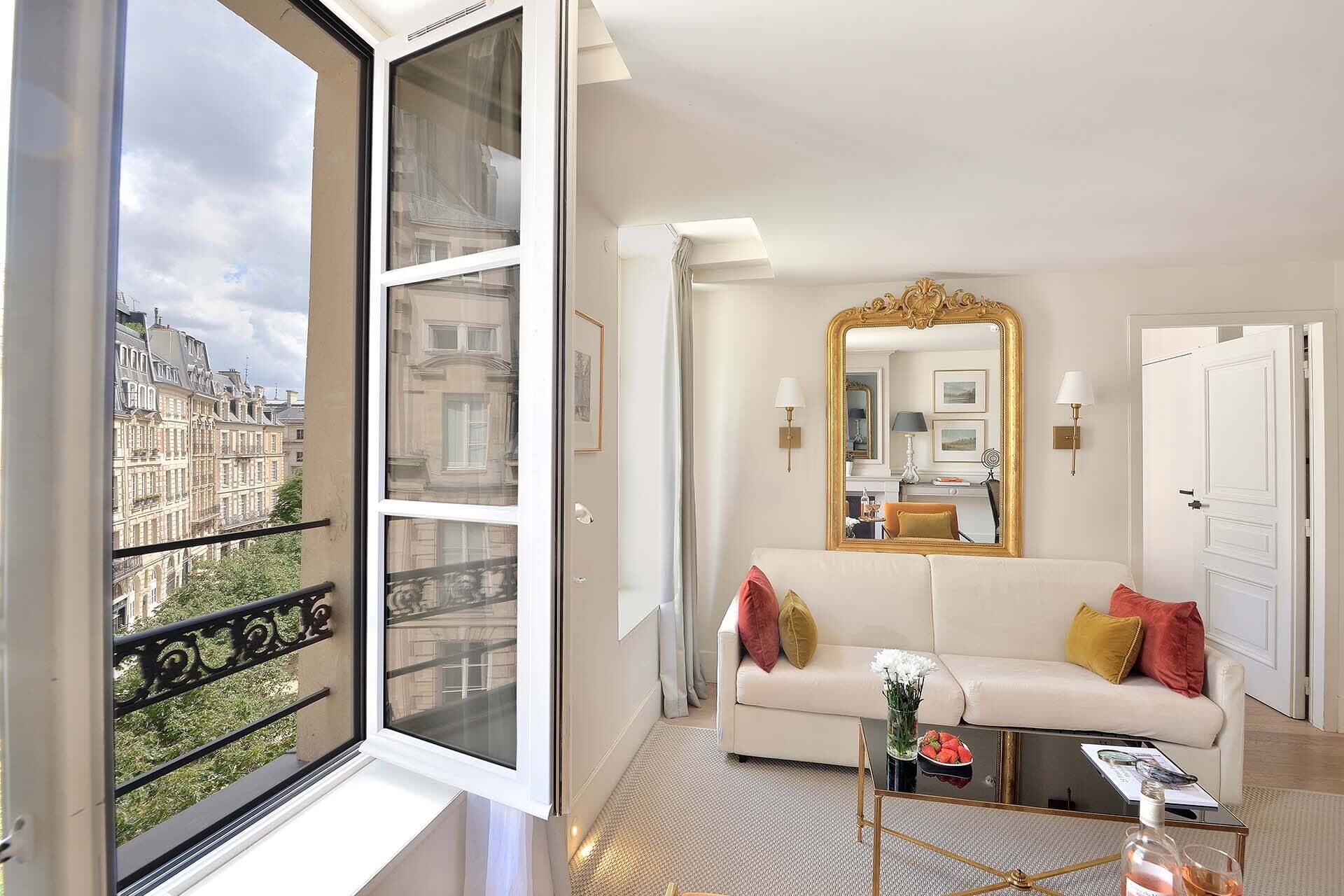A modern hotel room with a large window, a white sofa, a gold-framed mirror, and a small coffee table.