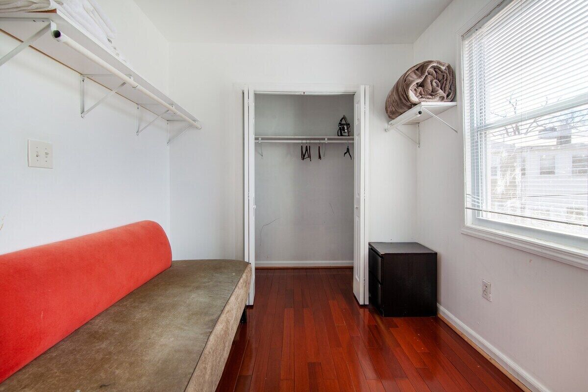 Ein Zimmer mit einem Bett, einem Kleiderschrank mit Kleidung, einem Heizkörper und einem Fenster mit Jalousien.