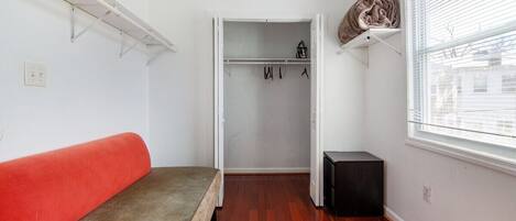 Ein Zimmer mit einem Bett, einem Kleiderschrank mit Kleidung, einem Heizkörper und einem Fenster mit Jalousien.