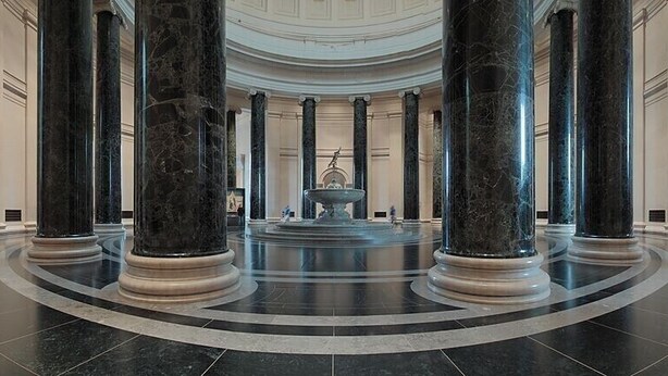 Un vaste hall d’entrée orné de grandes colonnes en marbre, doté d’une fontaine centrale et d’un plancher aux lignes courbes.