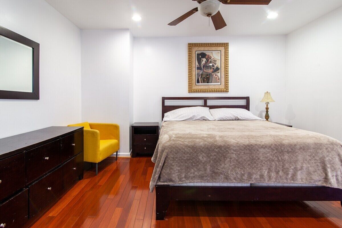 A bedroom with a bed, a yellow chair, a dark wooden dresser, a ceiling fan, and a framed picture on the wall.