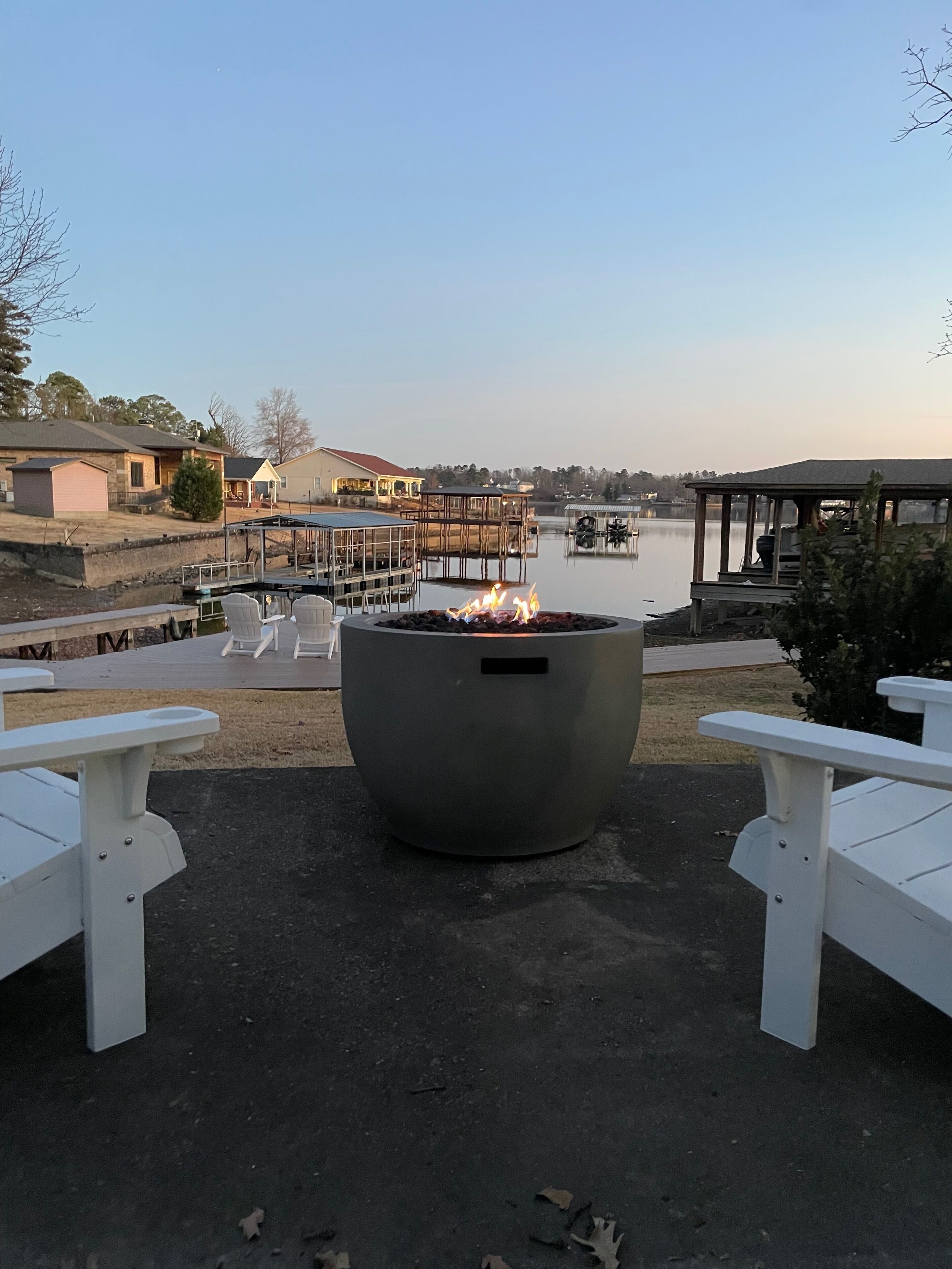 A fire pit in the center with white Adirondack chairs on either side, overlooking a calm lake with docked boats and a gazebo in the background.