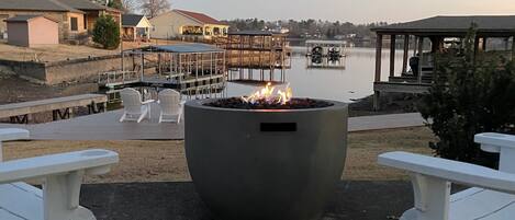 A fire pit in the center with white Adirondack chairs on either side, overlooking a calm lake with docked boats and a gazebo in the background.