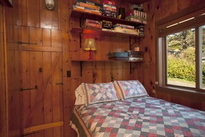 Una habitación con piso de madera, una cama, una ventana y estanterías llenas de libros y juegos.