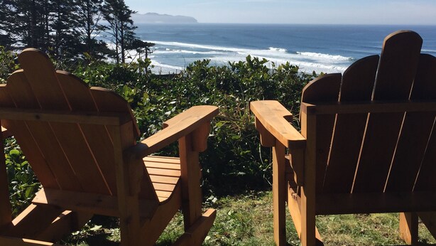 Two wooden Adirondack chairs facing a view of the ocean with waves and a distant island.