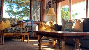 A cozy cabin interior with wooden walls and furniture, large windows offering a view of trees, and a bookshelf with books.